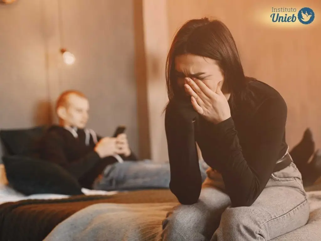 Mulher secando as lágrimas sentada na cama com marido ao fundo mexendo no celular, indicando uma situação de relacionamento conturbado