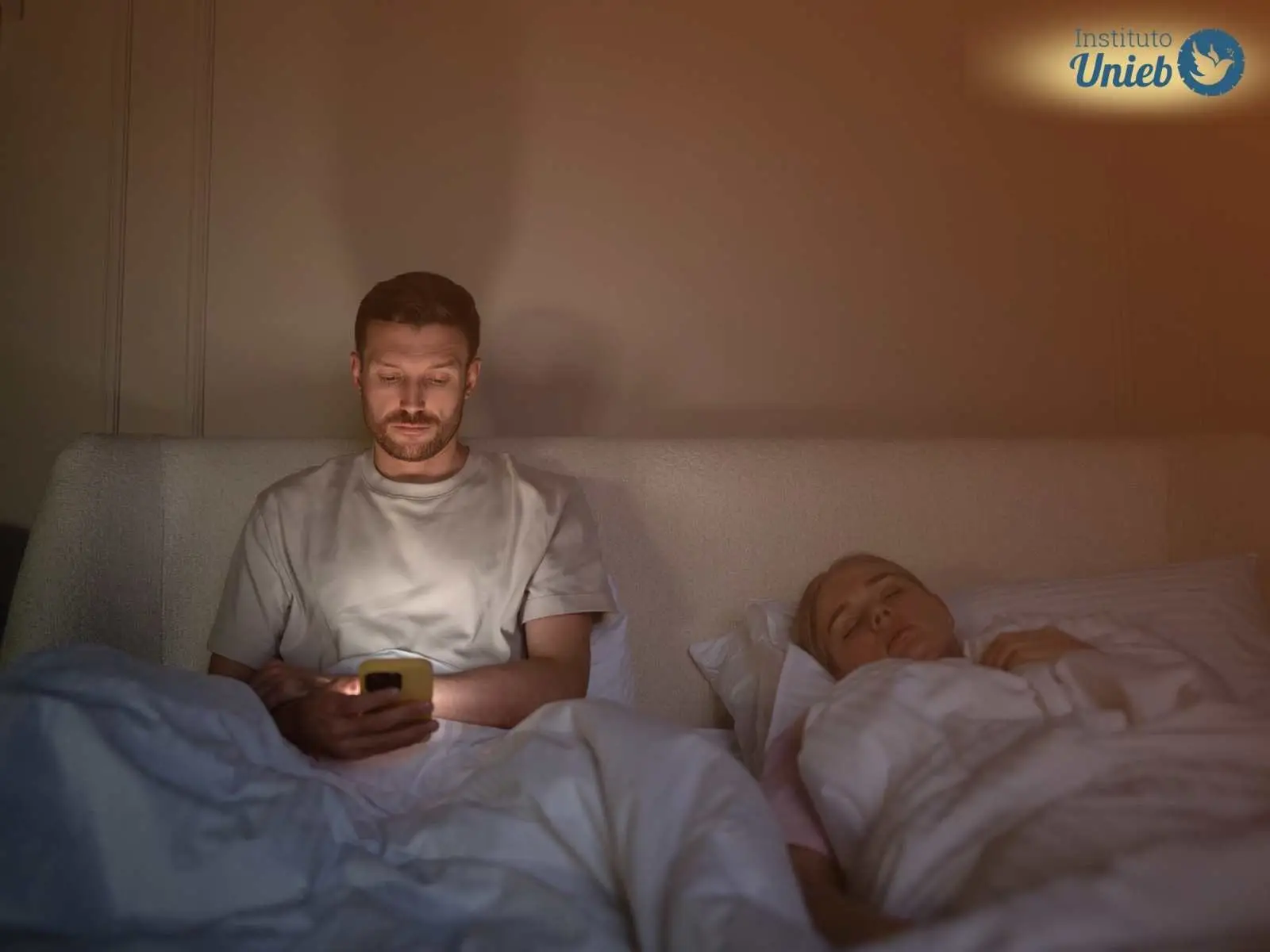 Homem deitado na cama mexendo no celular enquanto esposa dorme ao lado representando um marido que tem ciúmes da ex-esposa