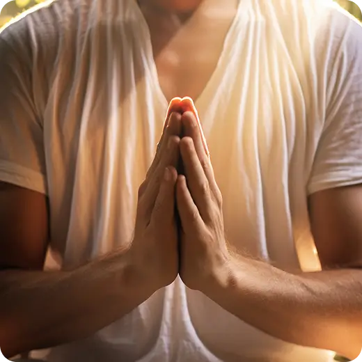 Pessoa com as palmas unidas em oração ou cumprimento, vestindo uma camisa branca e iluminada por uma luz quente e dourada.